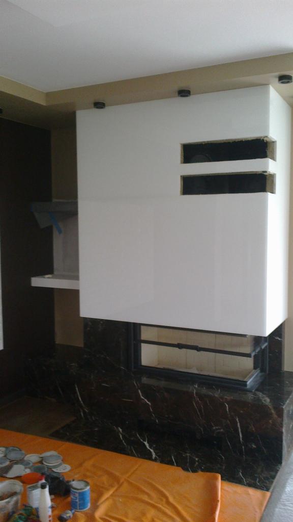 Biała obudowa kominka z dwoma poziomymi wnękami, czarnym marmurem u podstawy, pomarańczową folią ochronną na podłodze i narzędziami malarskimi obok.