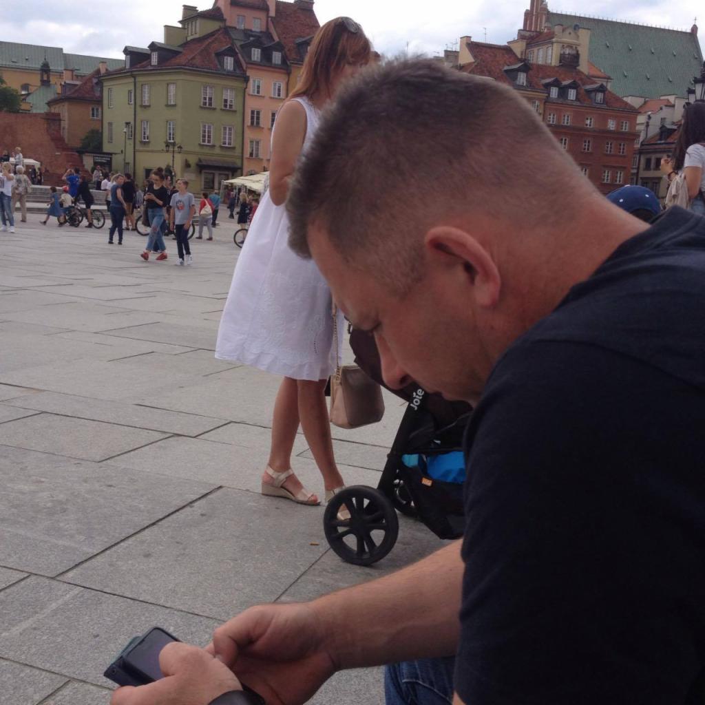 Mężczyzna siedzący na placu, przeglądający telefon, w tle kobieta w białej sukience i kolorowe kamienice Starego Miasta.