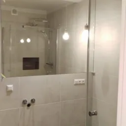 Nowoczesna łazienka z drewnopodobną podłogą, szklaną kabiną prysznicową i dużym lustrem, widoczne minimalistyczne oświetlenie sufitowe i wiszące lampy z żarówkami Edisona.