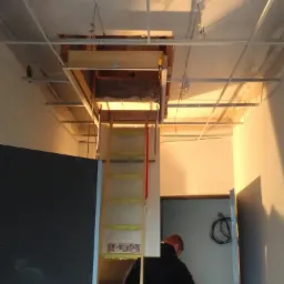 Montaż składanych schodów strychowych w suficie podwieszanym z widoczną konstrukcją nośną i częściowo ocieplonym poddaszem, pracownik na drabinie.