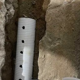 Krys - Biała rura drenażowa z otworami w wykopie ziemnym, widoczne fragmenty innych rur, ziemia o nierównej strukturze.