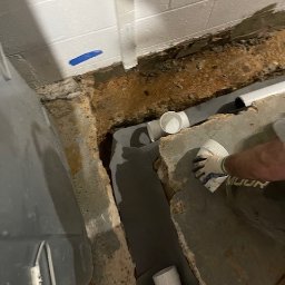 Krys - Instalacja kanalizacyjna w wykopie fundamentowym piwnicy z widoczną białą rurą PVC, szarym hydroizolacyjnym materiałem i fragmentem ściany z bloków betonowych z niebieską taśmą.