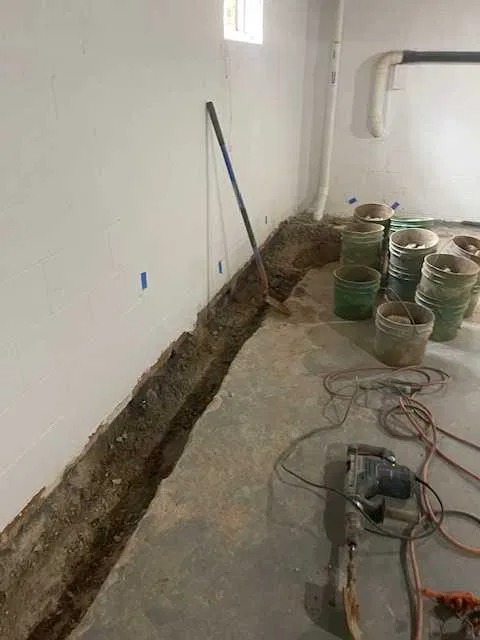 Wykopany rów wzdłuż ściany fundamentowej w piwnicy, przygotowany do izolacji przeciwwilgociowej, widoczne narzędzia i wiaderka z urobkiem.