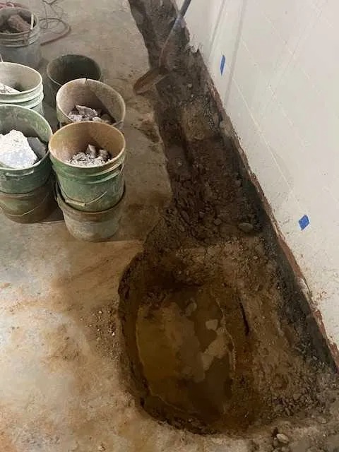 Wykop wzdłuż ściany fundamentowej, z widocznymi śladami wilgoci i stojącej wody w dole wykopu, obok ustawione wiaderka z gruzem i ziemią.