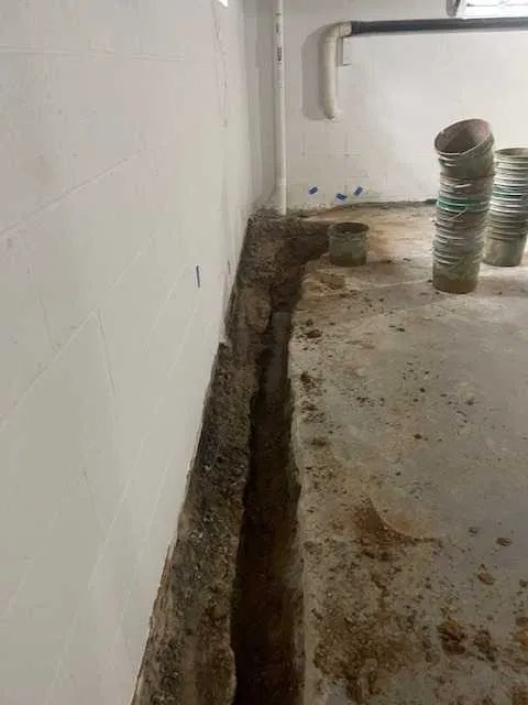 Wykop wzdłuż ściany fundamentowej w piwnicy, widoczne rury i wiadro, przygotowanie do izolacji lub drenażu.