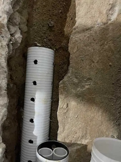 Biała rura drenażowa z otworami w wykopie ziemnym, widoczne fragmenty innych rur, ziemia o nierównej strukturze.