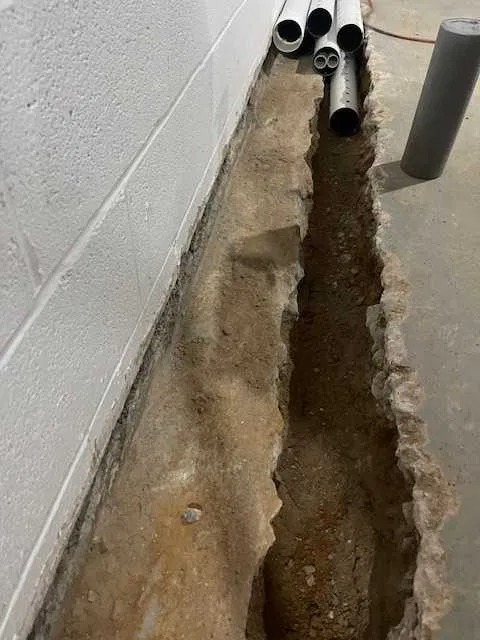 Wykop wzdłuż ściany fundamentowej w piwnicy, przygotowany pod instalację rur drenażowych, z widocznymi fragmentami gruzu i ziemi.