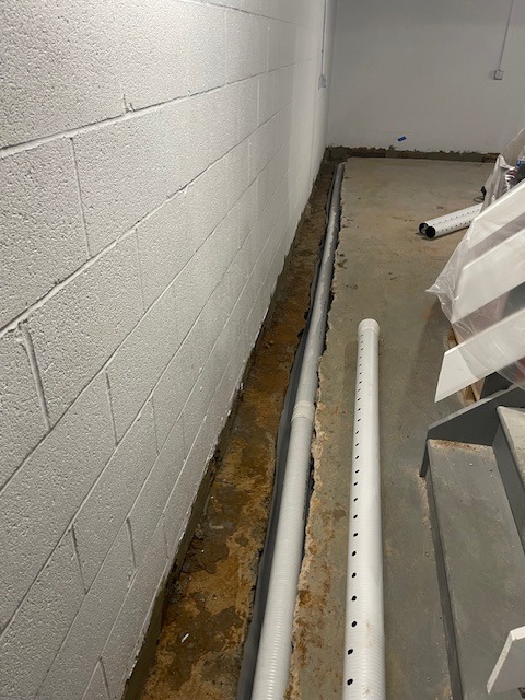 Drenaż wzdłuż ściany fundamentowej piwnicy, z widocznymi rurami drenażowymi z otworami i wykopem wypełnionym wodą, przygotowany do dalszych prac zabezpieczających przed wilgocią.