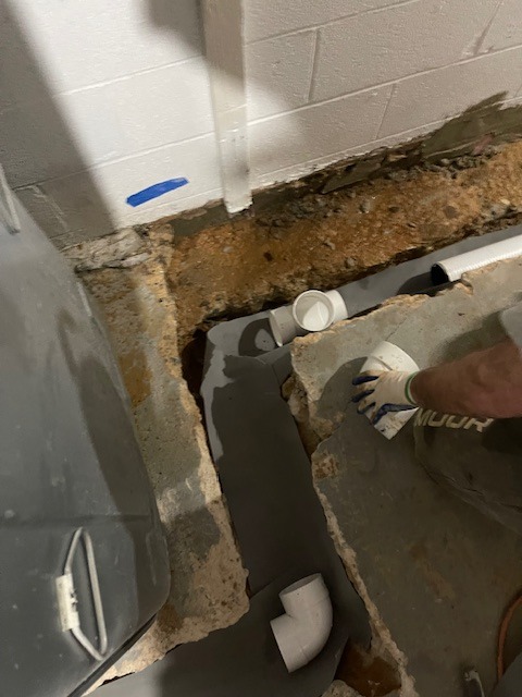 Instalacja kanalizacyjna w wykopie fundamentowym piwnicy z widoczną białą rurą PVC, szarym hydroizolacyjnym materiałem i fragmentem ściany z bloków betonowych z niebieską taśmą.