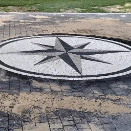 Kompozycja w kształcie rozety wkomponowana w nawierzchnię z kostki brukowej, z rozsypanym piaskiem na powierzchni i śladami wody po czyszczeniu.