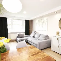 Jasny salon z szarą sofą narożną, drewnianym stołem z żółtymi kwiatami w wazonie, białą komodą z okrągłym lustrem i ścianą z cegły.