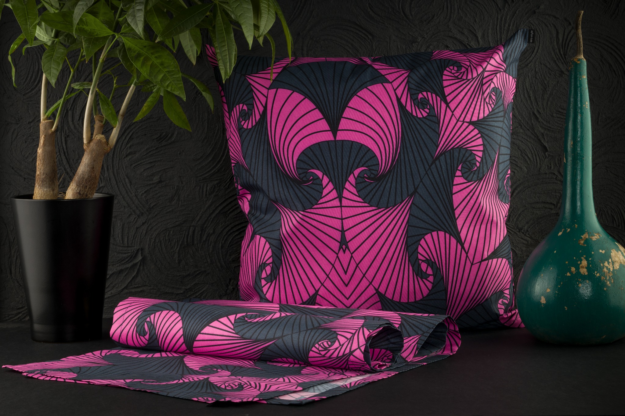 Poduszka dekoracyjna i zwinięty kawałek materiału z abstrakcyjnym wzorem w kolorach fuksji i grafitu na czarnym tle, obok doniczki z rośliną i zielonej, ozdobnej tykwy.