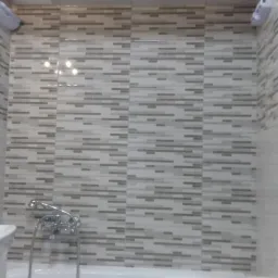 Wykończona łazienka z mozaiką z płytek w odcieniach szarości i beżu, umywalką z chromowaną baterią i lustrem w ciemnej ramie, z widoczną wanną i akcesoriami łazienkowymi.