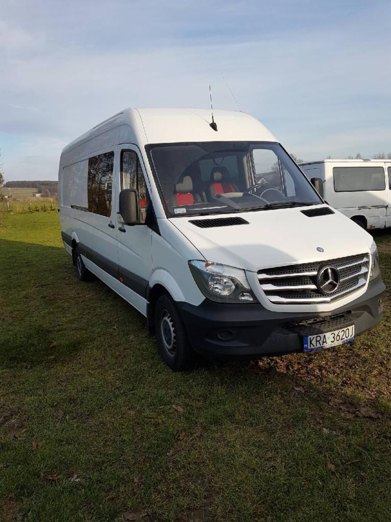 Biały bus Mercedes Sprinter z wysokim dachem, zaparkowany na trawiastym terenie, z widoczną tablicą rejestracyjną KRA 3620J i czerwonymi zagłówkami siedzeń przednich.