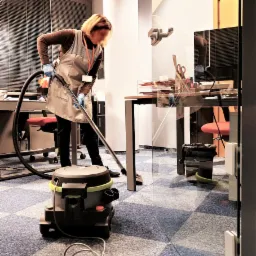 Kobieta w fartuchu i rękawicach odkurza wykładzinę biurową, widoczny odkurzacz przemysłowy i biurka z żaluzjami w tle.