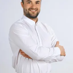 Portret uśmiechniętego mężczyzny w białej koszuli, z brodą i starannie ułożonymi włosami, pozującego z założonymi rękami na tle jasnego, neutralnego tła.