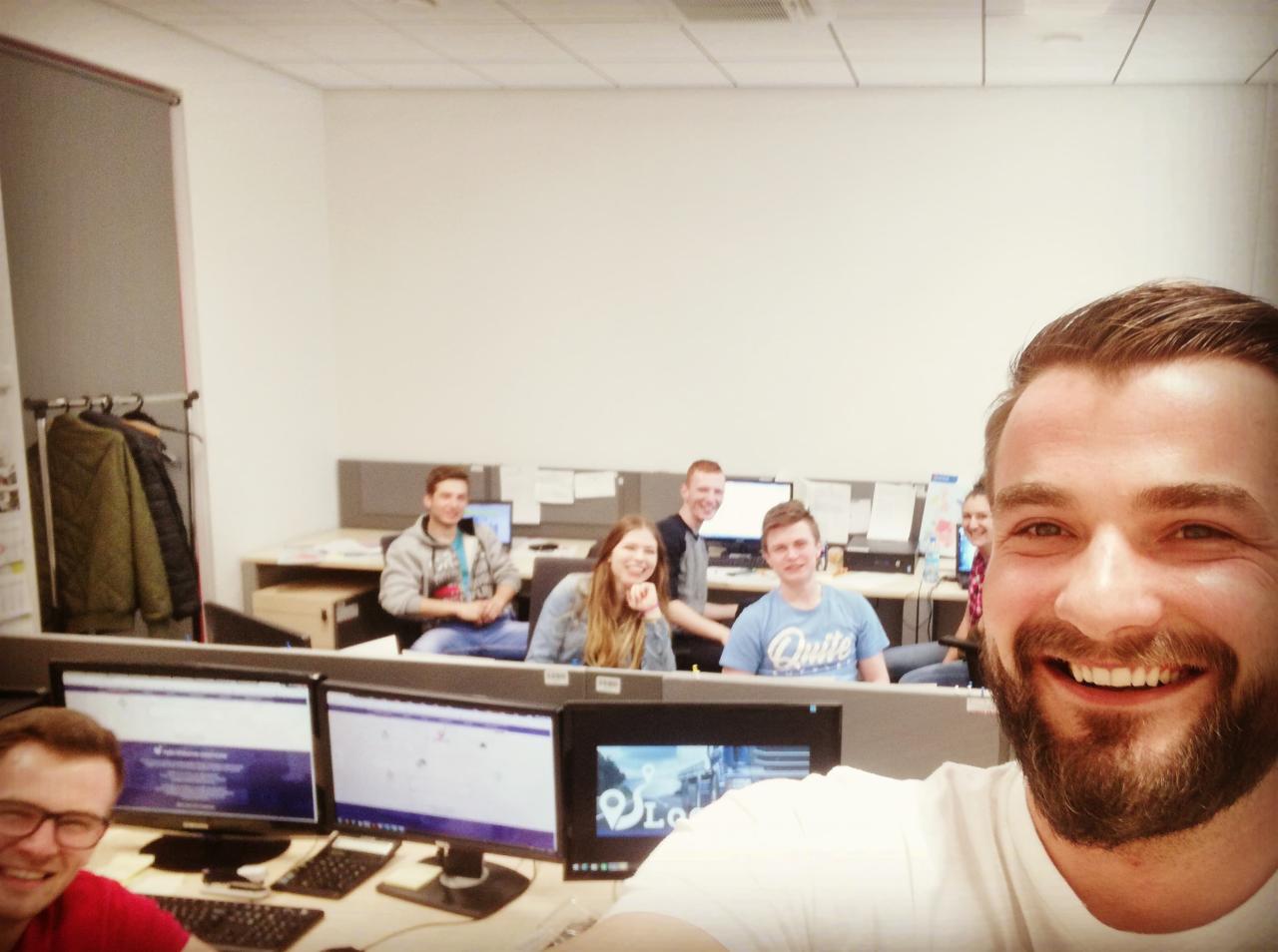 Uśmiechnięty mężczyzna robi selfie na tle biura z pracownikami siedzącymi przy komputerach, w tle wieszak z kurtkami.