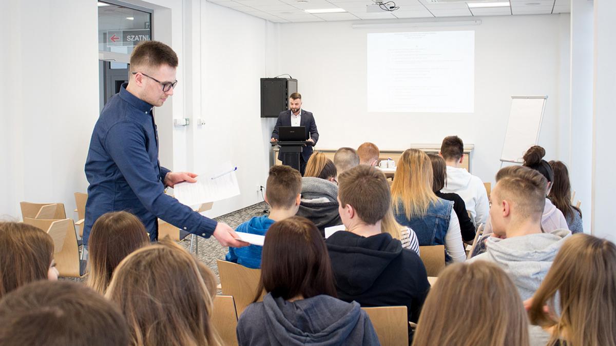 Sala konferencyjna podczas prezentacji: prelegent przy laptopie, mężczyzna w niebieskiej koszuli rozdaje dokumenty uczestnikom siedzącym na drewnianych krzesłach.