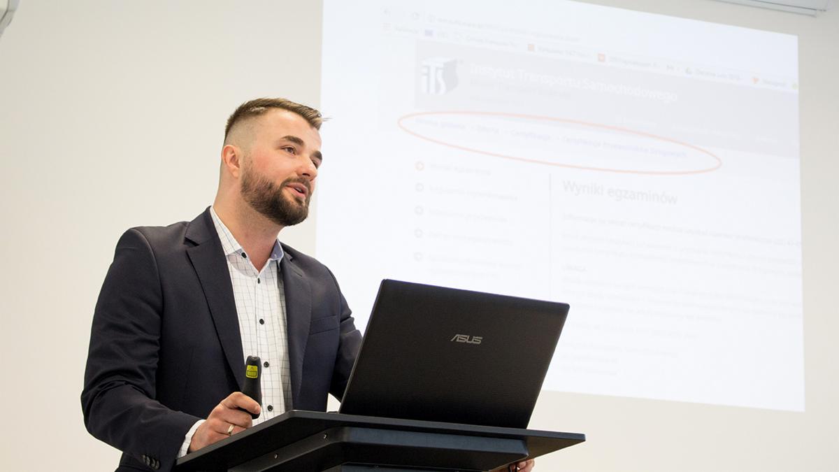 Mężczyzna w garniturze prezentuje wyniki egzaminów na stronie internetowej Instytutu Transportu Samochodowego, używając laptopa ASUS i wskaźnika.
