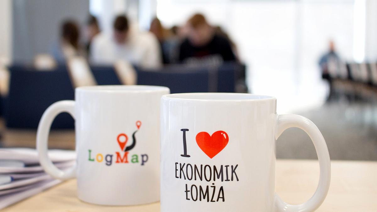 Dwa białe kubki z nadrukami na jasnym stole; jeden z logo LogMap, drugi z napisem 'I ❤️ EKONOMIK ŁOMŻA', w tle rozmyte postacie i krzesła.
