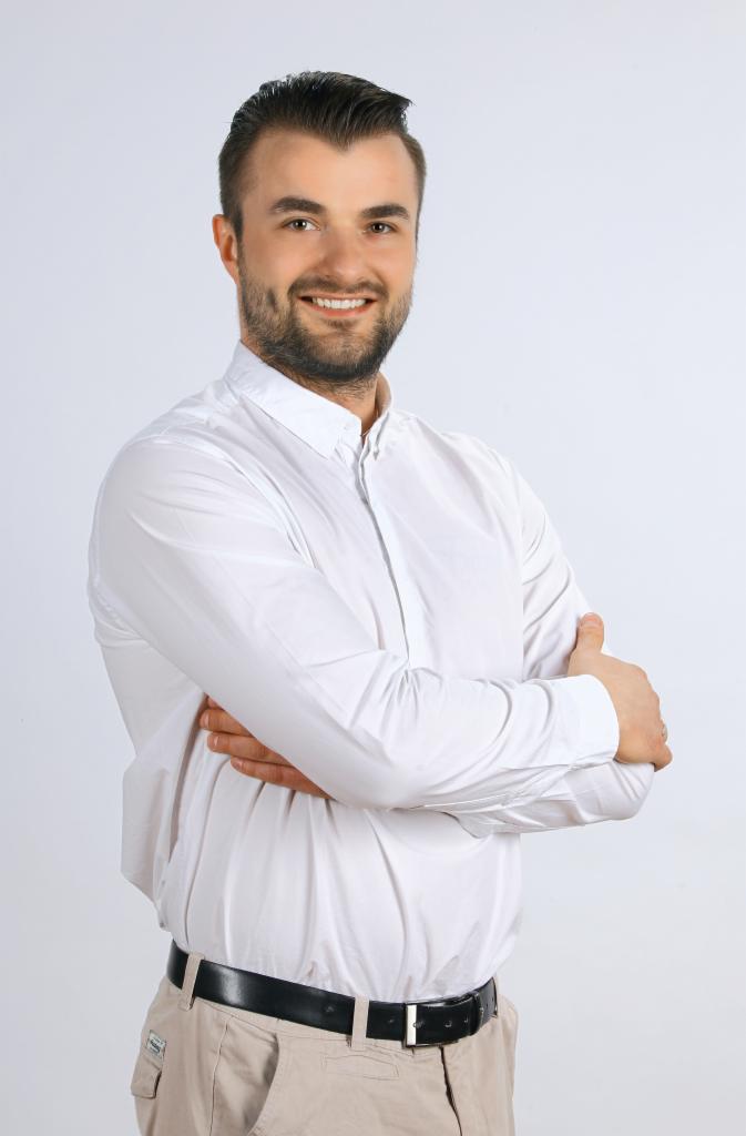 Portret uśmiechniętego mężczyzny w białej koszuli, z brodą i starannie ułożonymi włosami, pozującego z założonymi rękami na tle jasnego, neutralnego tła.