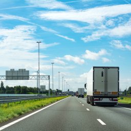 NOBELICA SPÓŁKA Z OGRANICZONĄ ODPOWIEDZIALNOŚCIĄ - Ciężarówka z białą naczepą na autostradzie w słoneczny dzień. Widoczne znaki drogowe i inne pojazdy. Perspektywa zza pojazdu, dynamiczne ujęcie w ruchu.