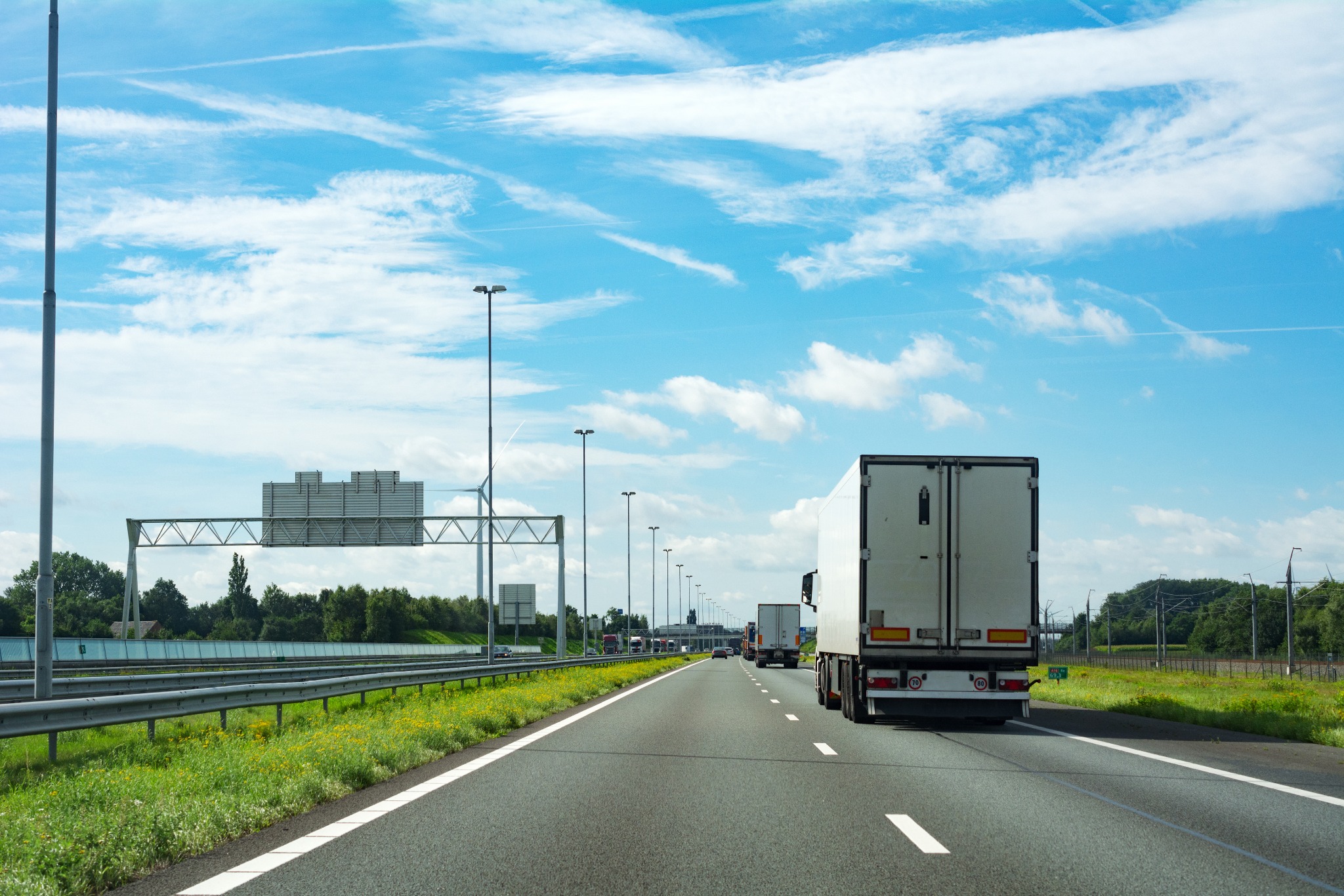 Ciężarówka z białą naczepą na autostradzie w słoneczny dzień. Widoczne znaki drogowe i inne pojazdy. Perspektywa zza pojazdu, dynamiczne ujęcie w ruchu.