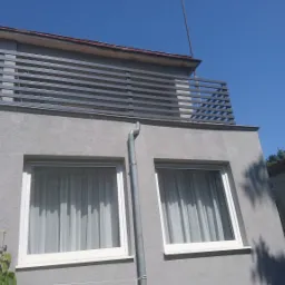 Szara elewacja budynku w Szczecinie z metalową balustradą balkonową o poziomym układzie szczebli, widok z dołu na błękitne niebo.