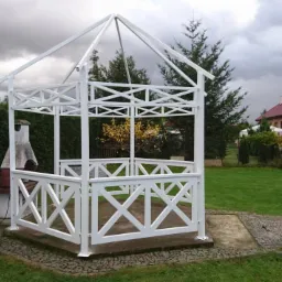 Biała altana ogrodowa z drewnianą konstrukcją kratową, na tle zielonego trawnika i zabudowań mieszkalnych pod pochmurnym niebem. Obok altany widoczny murowany grill.