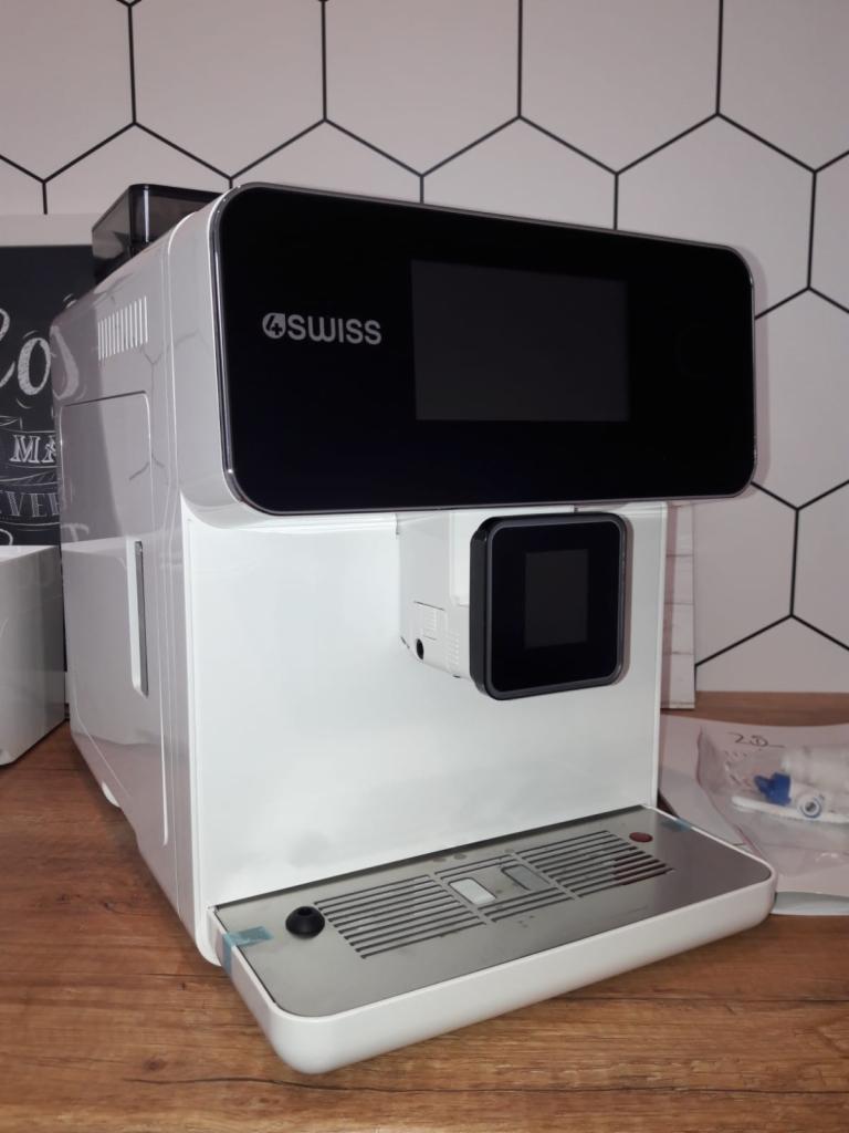 Biały, automatyczny ekspres do kawy marki Swiss z czarnym panelem dotykowym, stojący na drewnianym blacie, na tle ściany z heksagonalnymi płytkami.