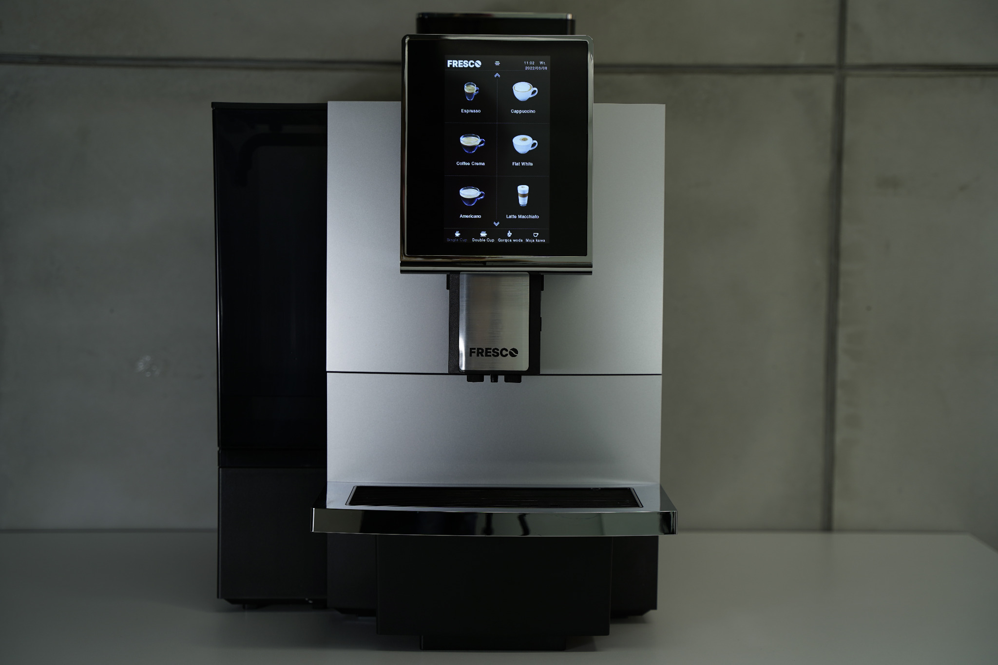 Automatyczny ekspres do kawy Fresco w kolorze srebrno-czarnym, z ekranem dotykowym wyświetlającym menu napojów, na tle betonowej ściany.