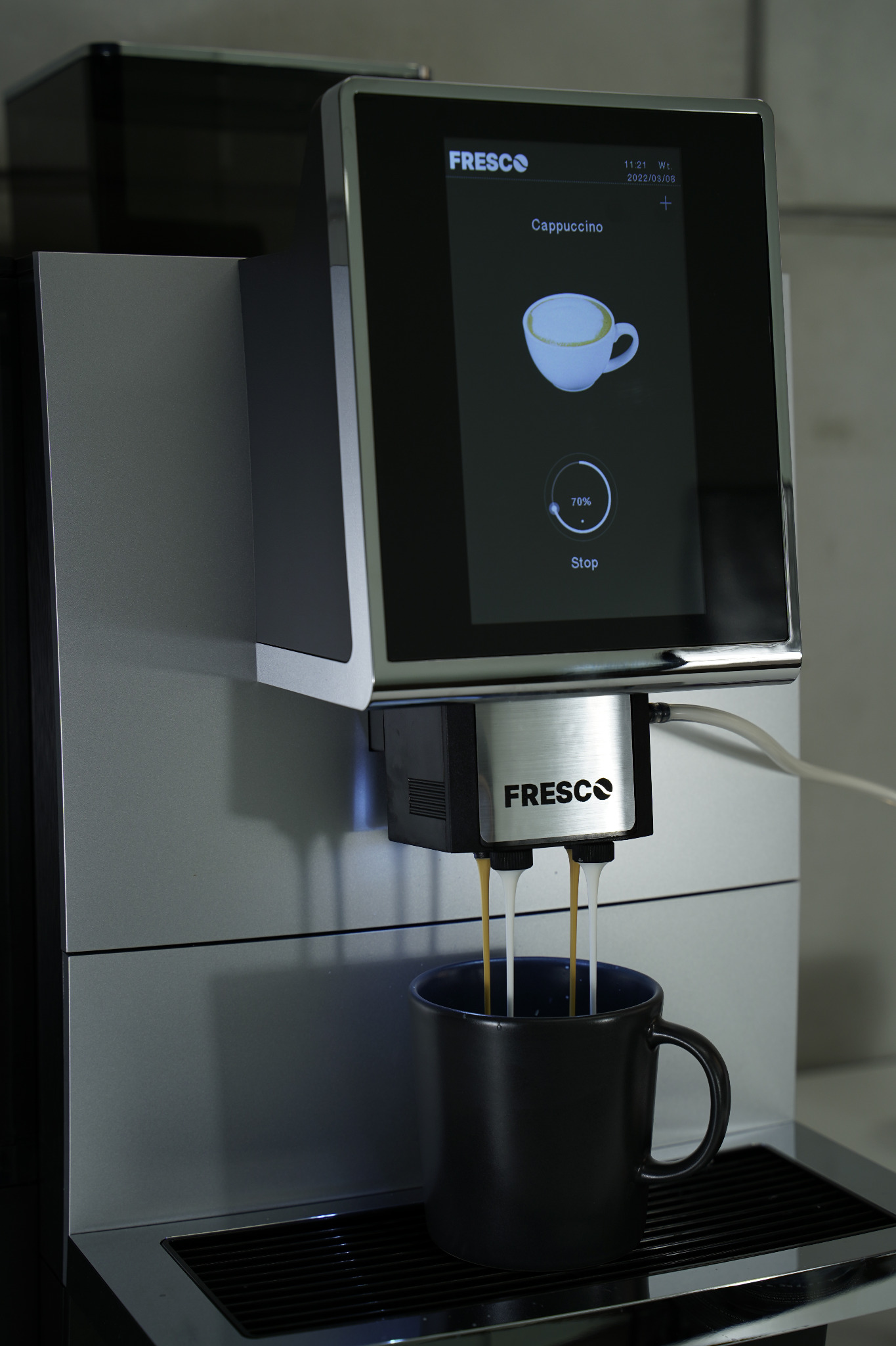 Automatyczny ekspres do kawy Fresco w trakcie przygotowywania cappuccino do czarnego kubka. Na ekranie dotykowym widoczny proces parzenia.