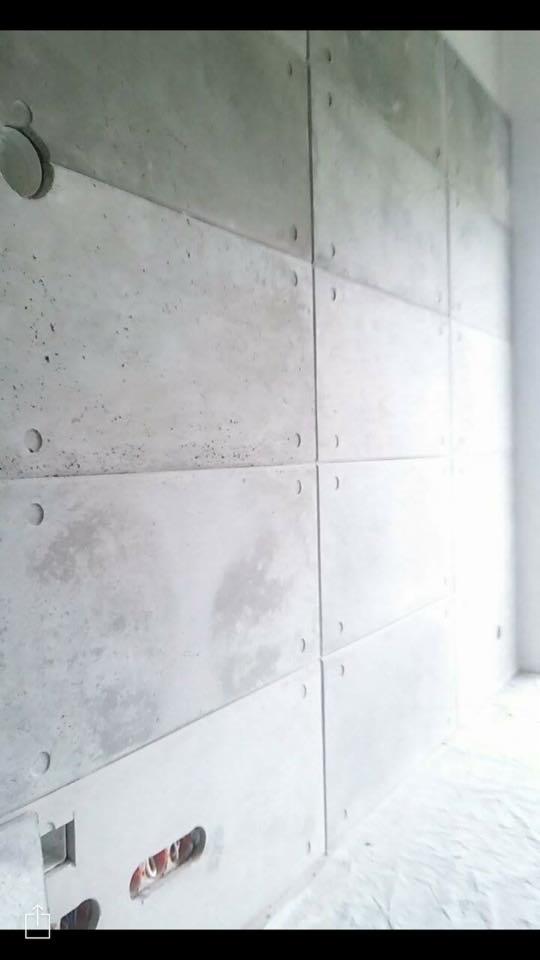 Ściana wykończona panelami z betonu architektonicznego z widocznymi otworami montażowymi i puszką elektryczną, zabezpieczona folią malarską.