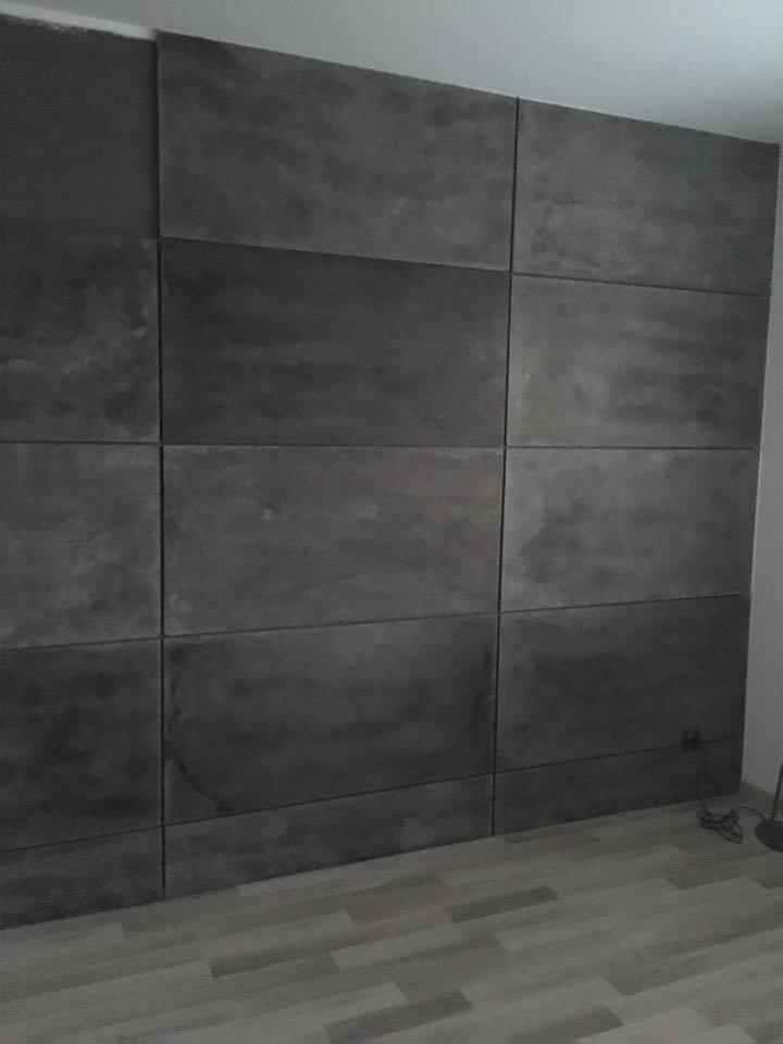 Ściana wyłożona ciemnoszarymi prefabrykatami betonowymi o nieregularnej fakturze, widoczne łączenia między płytami, szare panele podłogowe.