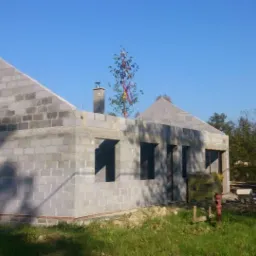 Surowy stan domu z szarego bloczka betonowego, widoczne otwory okienne i drzwiowe, w tle drzewa i samochód osobowy, na dachu komin.