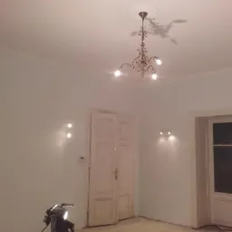 Wnętrze pokoju z świeżo pomalowanymi ścianami w odcieniach bieli, widoczne drzwi wewnętrzne i ozdobny żyrandol sufitowy. Na ścianach zamontowane kinkiety. W rogu kadru widoczny statyw oświetleniowy.