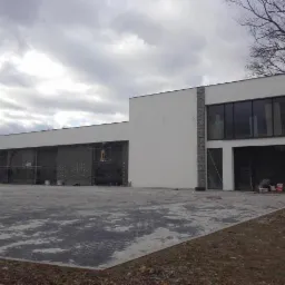 Nowoczesny budynek w trakcie ocieplania elewacji, widoczne rusztowania i materiały budowlane, szara kostka brukowa na podjeździe, pochmurne niebo.