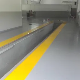 Szara posadzka przemysłowa z żółtymi pasami oznaczającymi kanał warsztatowy, widoczne oświetlenie w kanale i puszki z farbą w tle.