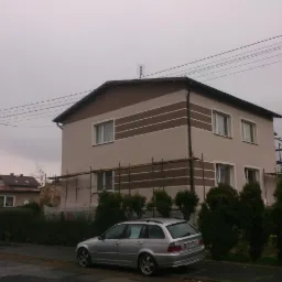 Dwupiętrowy dom z beżową elewacją i brązowymi pasami dekoracyjnymi, częściowo zakryty rusztowaniami, srebrny samochód kombi parkuje na ulicy przed domem.