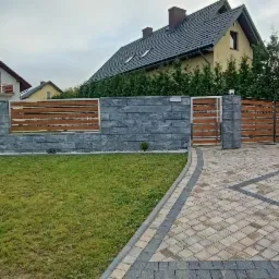 Nowoczesne ogrodzenie posesji z szarego kamienia łupanego i poziomych drewnianych paneli, przesuwna brama wjazdowa z drewna, kostka brukowa na podjeździe, domy w tle.