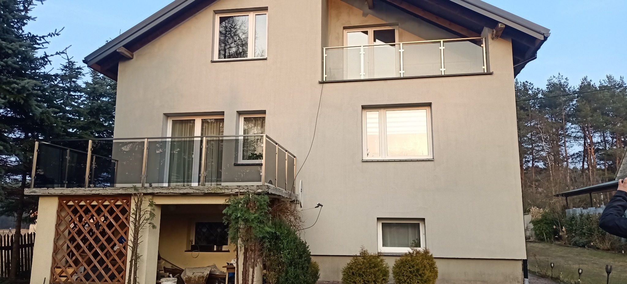 Dwukondygnacyjny dom z szarą elewacją i szklanymi balustradami balkonowymi, otoczony zielenią, widoczny fragment osoby w prawym dolnym rogu.