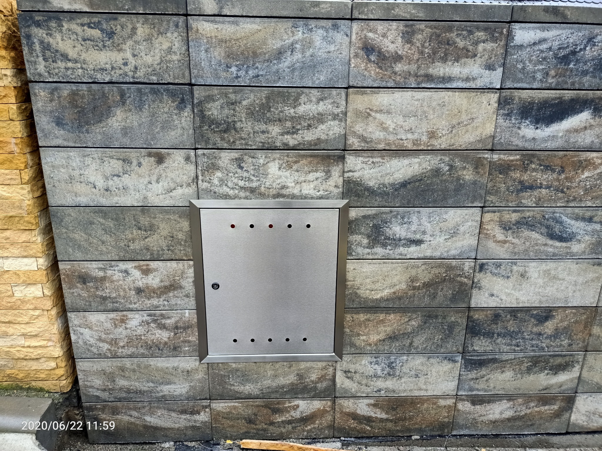 Metalowa skrzynka na gaz lub licznik, wbudowana w ścianę z szarych, cieniowanych bloczków betonowych, z widocznym niewielkim zamkiem i otworami wentylacyjnymi.