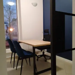 Nowoczesne biuro: jasny stół z czarnymi nogami, dwie granatowe krzesła, obrotowe krzesło biurowe z wysokim oparciem oraz widok na ulicę przez okno.