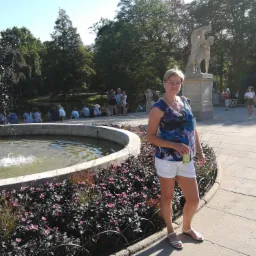 Kobieta w letnim stroju pozuje na tle fontanny i rzeźby w parku, trzymając napój w ręku.