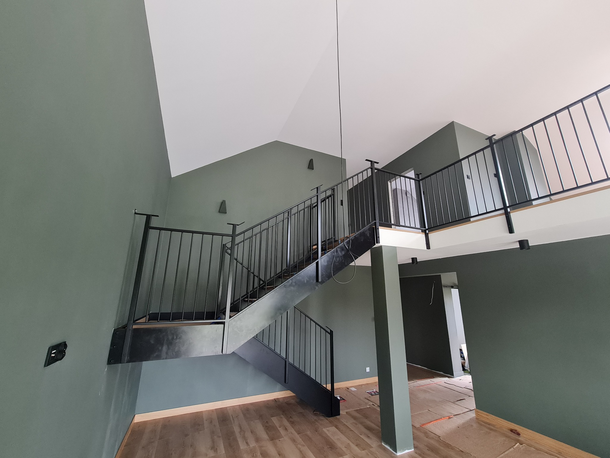 Nowoczesne schody metalowe z czarną balustradą w surowym wnętrzu z zielonymi ścianami i drewnopodobną podłogą, widok z dołu.