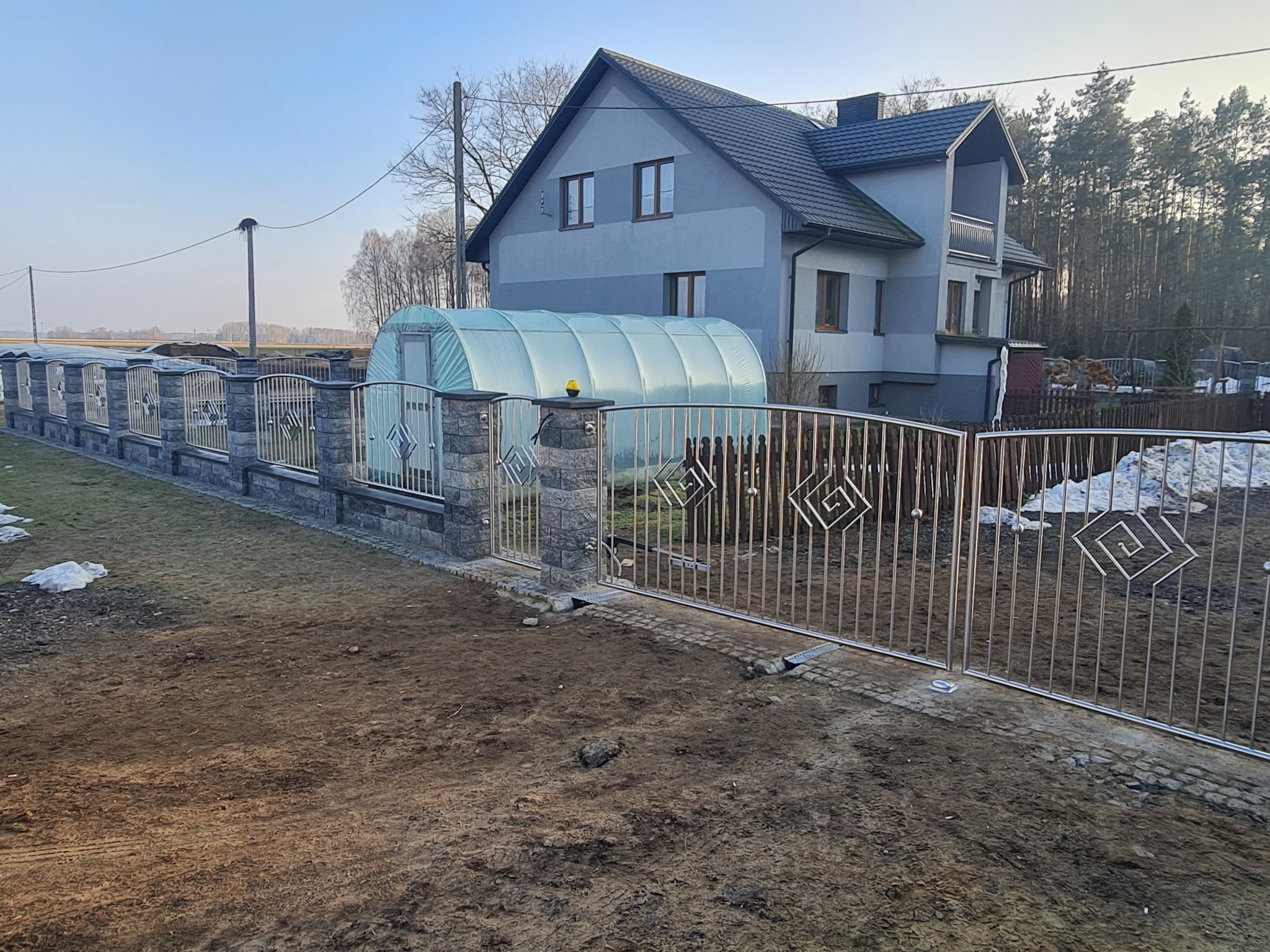 Nowoczesne ogrodzenie posesji ze stali nierdzewnej z elementami dekoracyjnymi w kształcie spiralnych kwadratów, na tle domu z szarą elewacją i szklarni.
