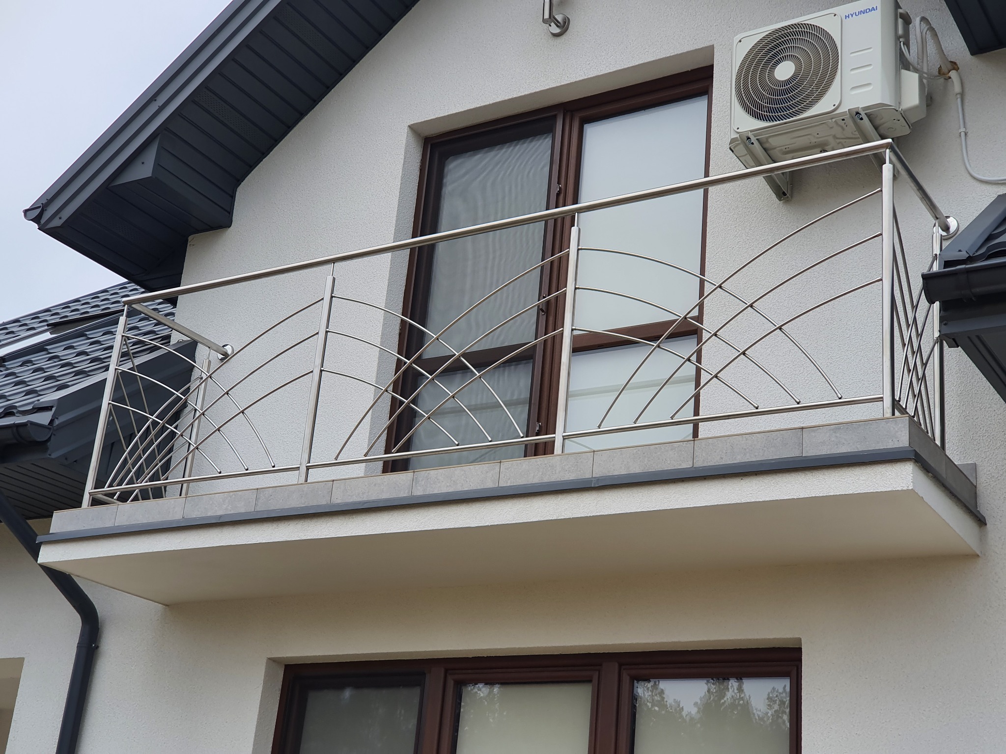 Balkon z nowoczesną balustradą ze stali nierdzewnej o dekoracyjnym, łukowatym wzorze, zamontowaną na zewnątrz budynku z klimatyzatorem na ścianie.