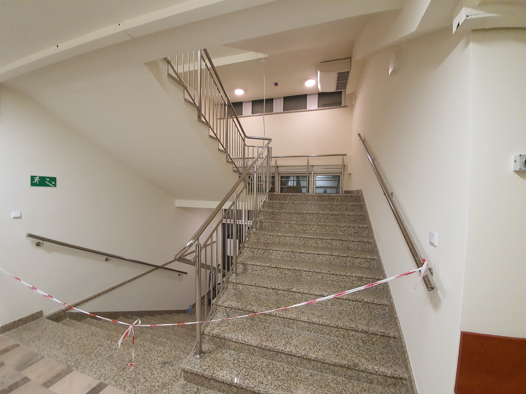 Wnętrze klatki schodowej z marmurowymi schodami i metalową balustradą, zabezpieczone taśmą ostrzegawczą.