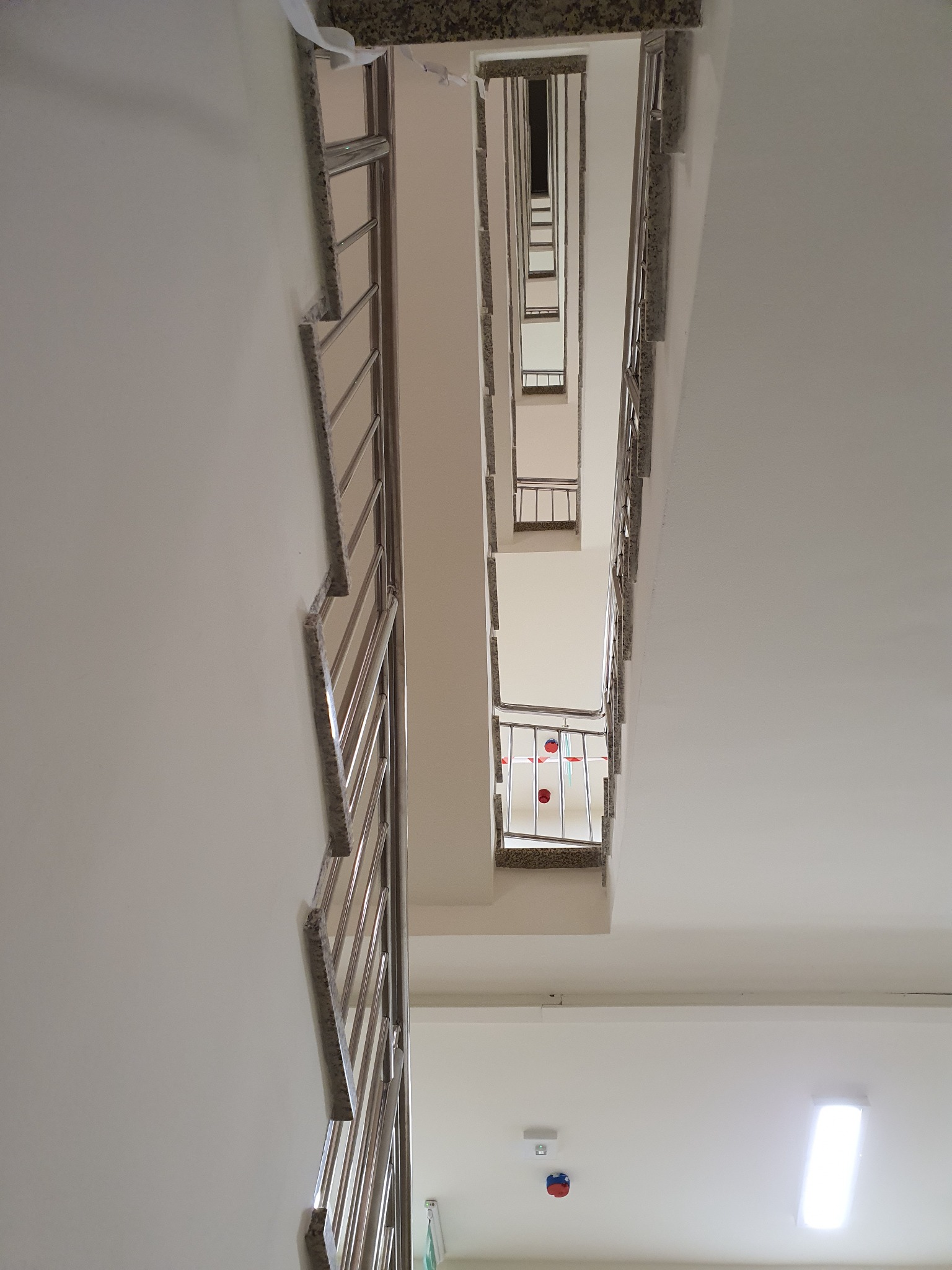 Widok z dołu na klatkę schodową z metalowymi balustradami, marmurowymi stopniami i białymi ścianami, perspektywa podkreśla pionowy układ schodów.