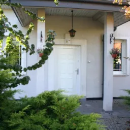 Wejście do domu z białymi drzwiami, oświetlone wiszącą lampą, z roślinami w doniczkach po bokach i zielenią na pierwszym planie.